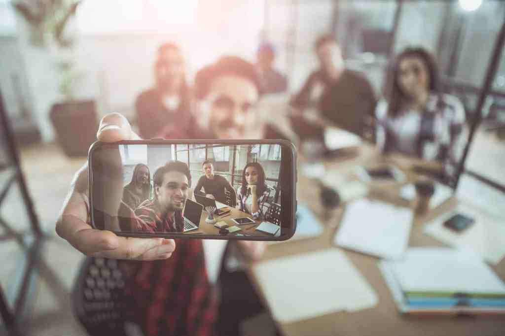 Selfie time. Close up of screen of modern smartphone in hand of cheerful guy. He is sitting at table in office with his members of team and taking picture of them. Focus on gadget with photo